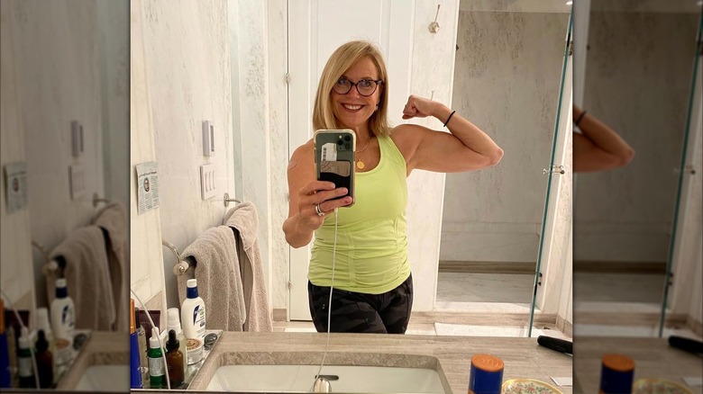 Katie Couric posing for a mirror selfie as she shows off bicep muscle. She is wearing a green workout tank and camouflage leggings.