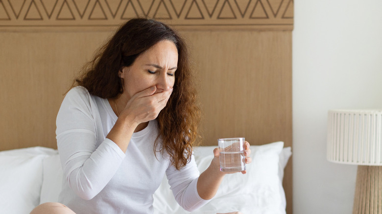 A woman feeling nauseated while holding a glass of water and sitting on her bed