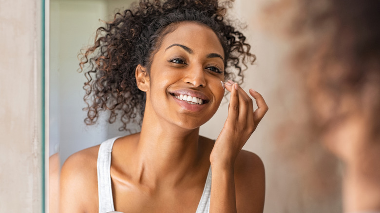 young woman smiling as she puts on moisturizer in the mirror