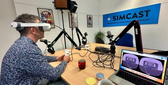 A man wearing a VR headset is sitting at a desk, and holding a single VR...